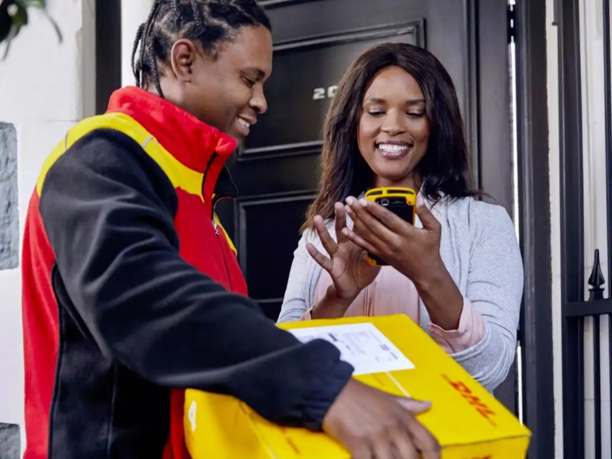 Imagem de um entregador da DHL sorrindo e entregando um pacote a uma mulher que está usando um smartphone. A interação ressalta a eficiência do serviço de entrega e o atendimento ao cliente.